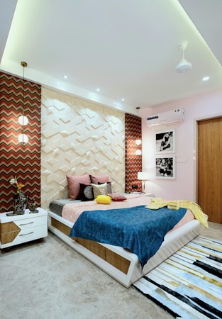 A modern bedroom with geometric wall panels and vibrant decor. The bed is adorned with colorful pillows and a blue blanket. Two pendant lights hang above each bedside table, enhancing the room's ambiance. Black and white photographs hang on the wall, and a textured rug lies on the floor.