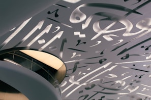 An architectural ceiling with intricate, abstract shapes and calligraphy designs. The ceiling features a mix of flowing curves and geometric forms, with light filtering through the spaces between the patterns. An elevated walkway with glass railings complements the modern aesthetic.