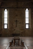 A dimly lit interior of a stone building features two tall, narrow stained-glass windows and a wooden cross mounted on the wall. The centerpiece includes a long wooden table with benches, set against the brick floor.