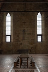 A dimly lit interior of a stone building features two tall, narrow stained-glass windows and a wooden cross mounted on the wall. The centerpiece includes a long wooden table with benches, set against the brick floor.