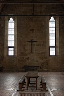 A dimly lit interior of a stone building features two tall, narrow stained-glass windows and a wooden cross mounted on the wall. The centerpiece includes a long wooden table with benches, set against the brick floor.
