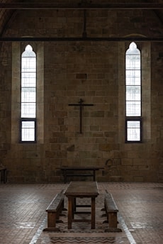 A dimly lit interior of a stone building features two tall, narrow stained-glass windows and a wooden cross mounted on the wall. The centerpiece includes a long wooden table with benches, set against the brick floor.