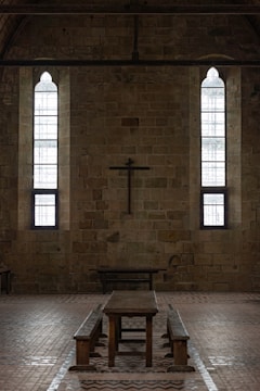 A dimly lit interior of a stone building features two tall, narrow stained-glass windows and a wooden cross mounted on the wall. The centerpiece includes a long wooden table with benches, set against the brick floor.