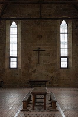 A dimly lit interior of a stone building features two tall, narrow stained-glass windows and a wooden cross mounted on the wall. The centerpiece includes a long wooden table with benches, set against the brick floor.