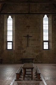 A dimly lit interior of a stone building features two tall, narrow stained-glass windows and a wooden cross mounted on the wall. The centerpiece includes a long wooden table with benches, set against the brick floor.