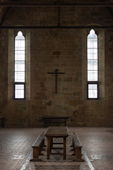 A dimly lit interior of a stone building features two tall, narrow stained-glass windows and a wooden cross mounted on the wall. The centerpiece includes a long wooden table with benches, set against the brick floor.