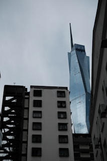 A futuristic skyscraper with angular, reflective surfaces towers above older, worn apartment buildings with multiple windows. The sky is overcast, casting a somber light over the scene.