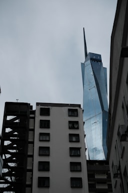 A futuristic skyscraper with angular, reflective surfaces towers above older, worn apartment buildings with multiple windows. The sky is overcast, casting a somber light over the scene.