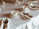 A neatly set dining table featuring white porcelain tea cups with saucers, folded napkins with silverware, and clear wine glasses. The arrangement is elegant and organized, suggesting a formal dining experience.