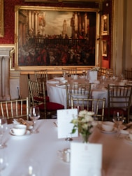 A formal dining room with round tables covered in white tablecloths, set with fine china, glasses, and menus. Elegant gold chairs surround the tables, and a large, ornate painting hangs on a rich, red-patterned wall. The room exudes a sense of classic elegance with its sophisticated decor.
