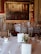 A formal dining room with round tables covered in white tablecloths, set with fine china, glasses, and menus. Elegant gold chairs surround the tables, and a large, ornate painting hangs on a rich, red-patterned wall. The room exudes a sense of classic elegance with its sophisticated decor.