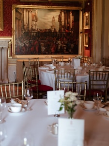 A formal dining room with round tables covered in white tablecloths, set with fine china, glasses, and menus. Elegant gold chairs surround the tables, and a large, ornate painting hangs on a rich, red-patterned wall. The room exudes a sense of classic elegance with its sophisticated decor.