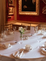 An elegantly set round dining table with white tablecloths, neatly arranged teacups, saucers, and silverware surrounds a central vase with white flowers. There are several wine glasses placed upside down. The background features rich red walls adorned with ornate framed paintings, contributing to a classic and sophisticated ambiance.