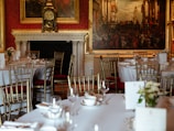 A luxurious dining room setting featuring elegantly arranged tables with white tablecloths, gold-colored chairs, and neatly placed tableware. The background showcases a grand fireplace with an ornate clock and large classical paintings adorning the walls.