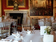 A luxurious dining room setting featuring elegantly arranged tables with white tablecloths, gold-colored chairs, and neatly placed tableware. The background showcases a grand fireplace with an ornate clock and large classical paintings adorning the walls.
