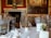 A luxurious dining room setting featuring elegantly arranged tables with white tablecloths, gold-colored chairs, and neatly placed tableware. The background showcases a grand fireplace with an ornate clock and large classical paintings adorning the walls.
