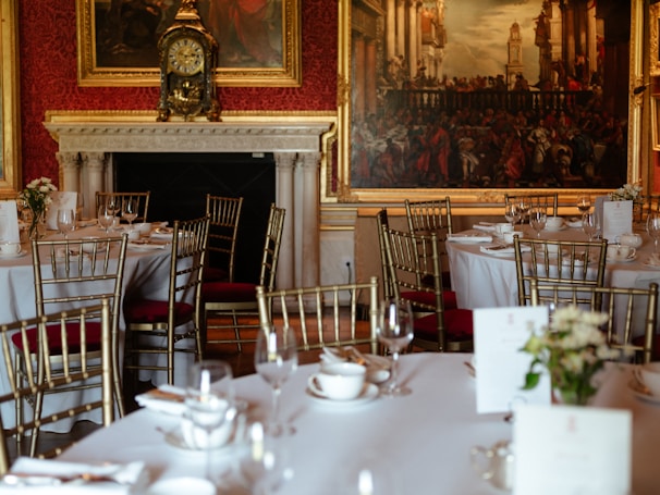 A luxurious dining room setting featuring elegantly arranged tables with white tablecloths, gold-colored chairs, and neatly placed tableware. The background showcases a grand fireplace with an ornate clock and large classical paintings adorning the walls.