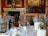 A luxurious dining room setting featuring elegantly arranged tables with white tablecloths, gold-colored chairs, and neatly placed tableware. The background showcases a grand fireplace with an ornate clock and large classical paintings adorning the walls.