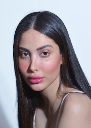 A person with long, straight dark hair and natural makeup, featuring pink lipstick and shimmering eyeshadow. The lighting highlights the facial features against a light background.
