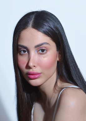 A person with long, straight dark hair and natural makeup, featuring pink lipstick and shimmering eyeshadow. The lighting highlights the facial features against a light background.