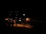 A nighttime street scene with dimly lit pathways and a prominent sign reading 'MUSSOORIE QUEEN'. There are people walking and a rickshaw with a passenger in the foreground. Streetlights illuminate the path slightly, while lights are visible from distant buildings on the hillside.