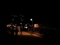 A nighttime street scene with dimly lit pathways and a prominent sign reading 'MUSSOORIE QUEEN'. There are people walking and a rickshaw with a passenger in the foreground. Streetlights illuminate the path slightly, while lights are visible from distant buildings on the hillside.
