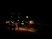 A nighttime street scene with dimly lit pathways and a prominent sign reading 'MUSSOORIE QUEEN'. There are people walking and a rickshaw with a passenger in the foreground. Streetlights illuminate the path slightly, while lights are visible from distant buildings on the hillside.