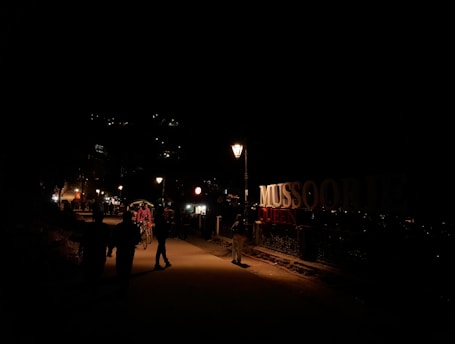 A nighttime street scene with dimly lit pathways and a prominent sign reading 'MUSSOORIE QUEEN'. There are people walking and a rickshaw with a passenger in the foreground. Streetlights illuminate the path slightly, while lights are visible from distant buildings on the hillside.