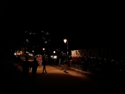A nighttime street scene with dimly lit pathways and a prominent sign reading 'MUSSOORIE QUEEN'. There are people walking and a rickshaw with a passenger in the foreground. Streetlights illuminate the path slightly, while lights are visible from distant buildings on the hillside.