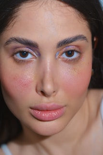 A close-up of a woman's face with shimmering makeup on her cheeks and eyelids. Her eyes are prominent with a blue tint on the eyelids and natural lips. The lighting highlights her glowing skin.