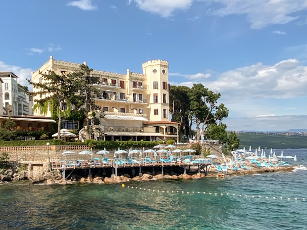 A large, elegant building with a castle-like tower located by the seaside. White and blue parasols and lounge chairs are arranged on a stone patio near the water. The landscape includes lush greenery and a few tall trees. The sea is calm, and the sky is partly cloudy, creating a picturesque coastal scene.