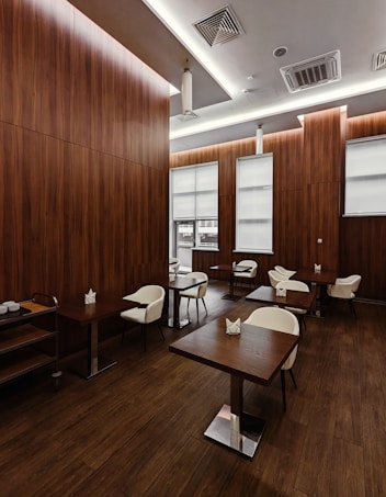 A modern and minimalist dining area features wooden walls and flooring that create a warm atmosphere. The room contains square wooden tables paired with white cushioned chairs arranged neatly. The ceiling design includes recessed lighting and air vents, contributing to a clean and sophisticated look. Large windows with blinds allow natural light to filter through.