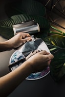 Hands are peeling a piece of metallic foil off a round object with colorful graphics. Large green leaves and a metallic roll of foil are placed on a dark surface in the background.