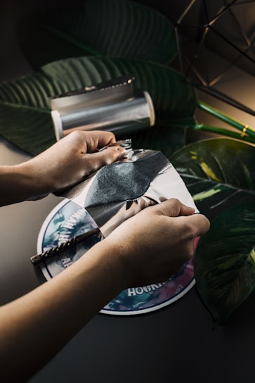 Hands are peeling a piece of metallic foil off a round object with colorful graphics. Large green leaves and a metallic roll of foil are placed on a dark surface in the background.