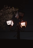 Three street lamps with lantern-like designs are illuminated against a dark night sky. The lamps emit a warm orange glow, creating a soft ambiance. Leaves from a nearby tree partially obscure the lamps, adding a natural element to the scene.