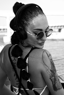 A woman with a braided ponytail and headphones, wearing sunglasses and displaying prominent tattoos on her shoulder and arm. Her skin is shimmering under the light, and she appears to be near a waterfront or pool setting.