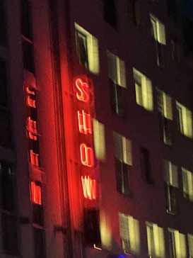 Neon red lights spell out 'SHOW' vertically on the side of a building at night. The building has multiple windows faintly illuminated from the inside.