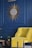 A stylish interior setup featuring a yellow chair with an ottoman, positioned against a deep blue wall. Above the chair is a decorative sunburst mirror with intricate golden rays. A small plant is placed on a round wooden side table, adding a touch of greenery to the scene.