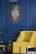 A stylish interior setup featuring a yellow chair with an ottoman, positioned against a deep blue wall. Above the chair is a decorative sunburst mirror with intricate golden rays. A small plant is placed on a round wooden side table, adding a touch of greenery to the scene.