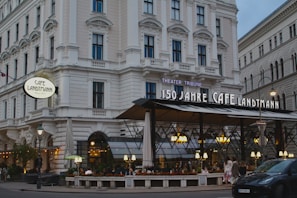 A large, elegant café with a classic architectural facade featuring multiple windows and decorative masonry. The café has an outdoor seating area with overhead lighting and several people dining. The signage indicates it's celebrating its 150th anniversary. Cars are parked along the adjacent street.