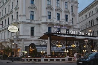 A large, elegant café with a classic architectural facade featuring multiple windows and decorative masonry. The café has an outdoor seating area with overhead lighting and several people dining. The signage indicates it's celebrating its 150th anniversary. Cars are parked along the adjacent street.