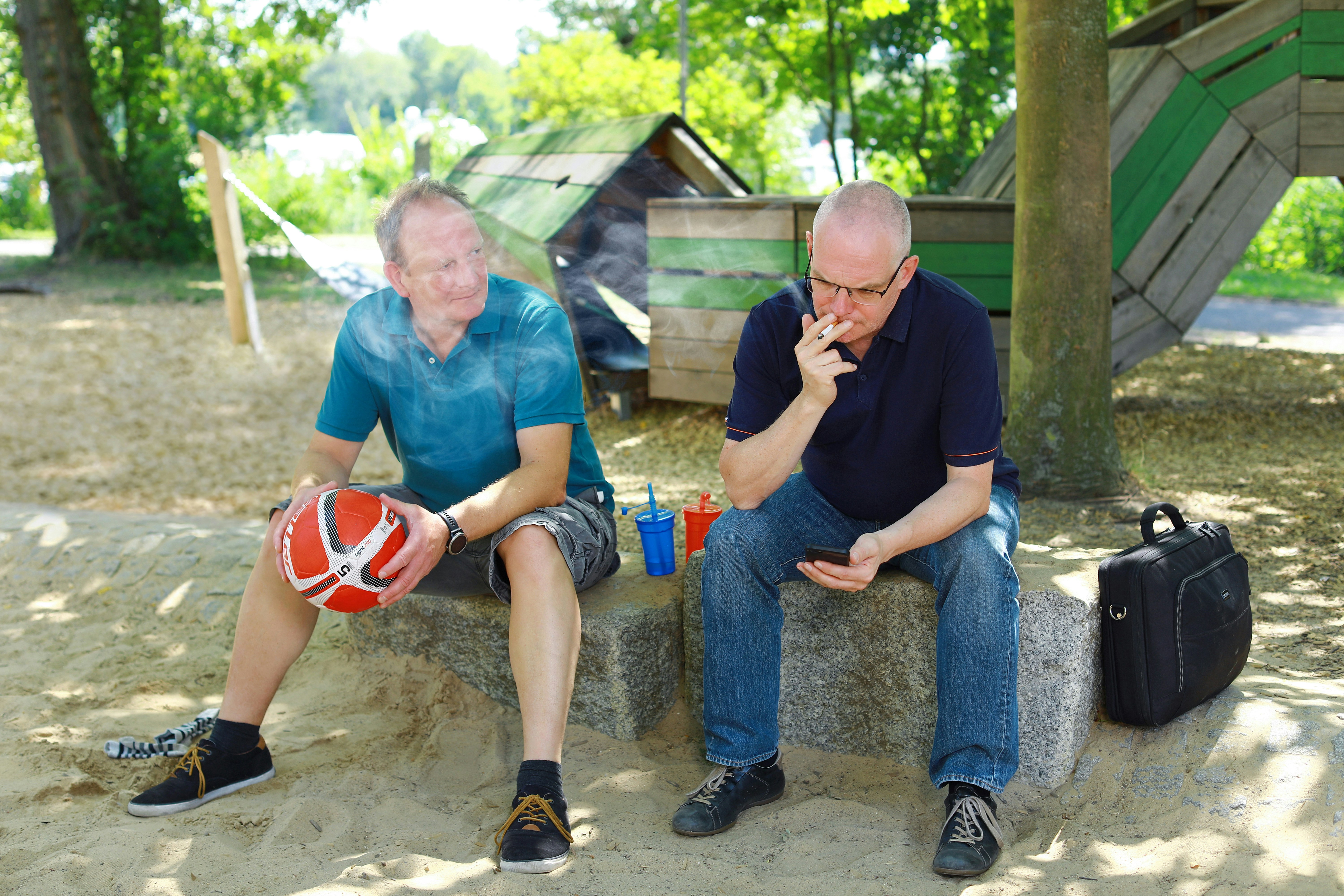 A men smokes a cigarette outside and disturbs his friend, an active, sporty mid age men, with his smoke - an appeal for a little more attention and awareness when smoking outdoors. Ein Mann raucht im Außenbereich eines Parks eine Zigarette und belästigt mit ihrem Rauch seinen Freund, einen sportlichen Mann - ein Aufruf zu etwas mehr Rücksicht und Achtsamkeit beim Rauchen im Freien.