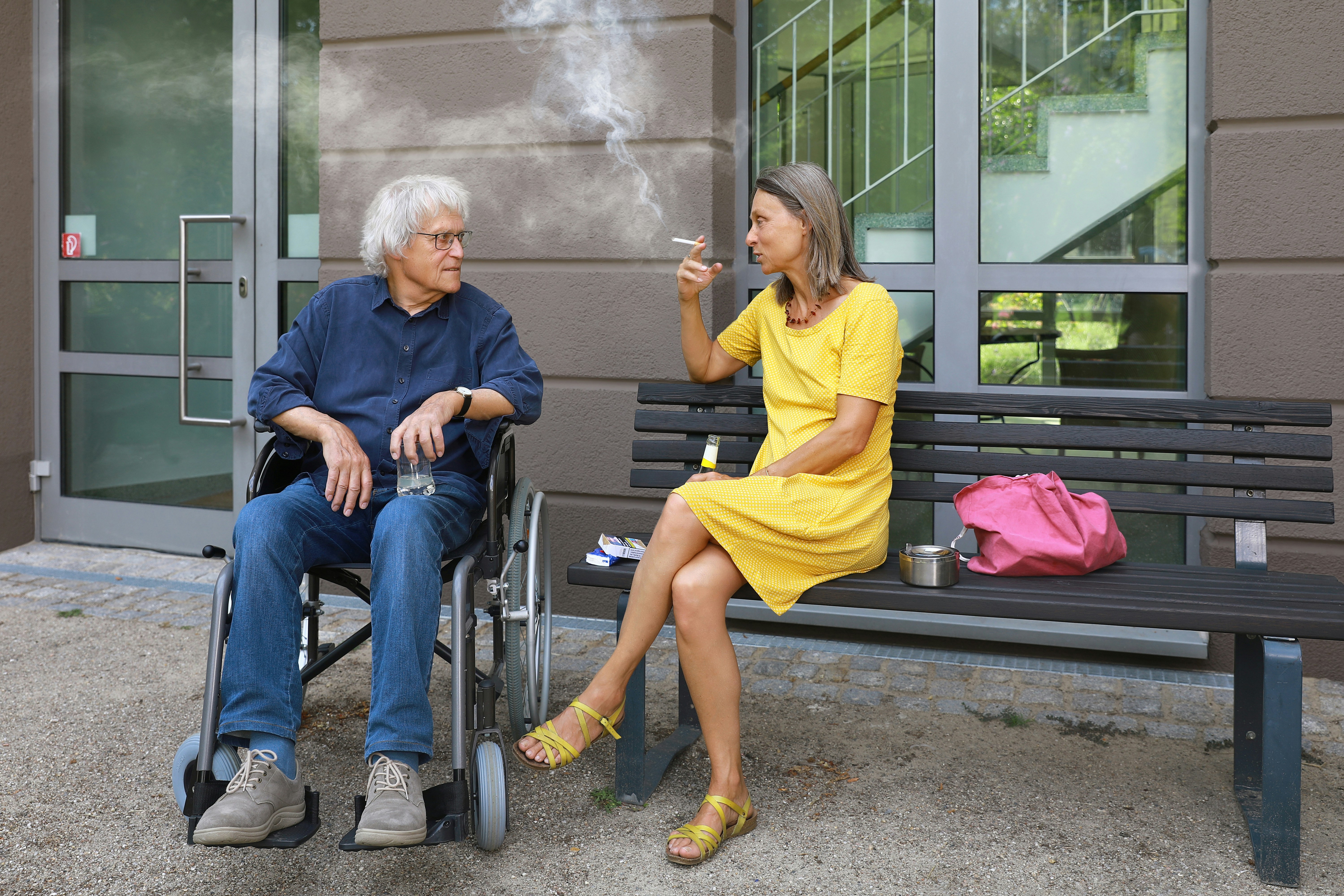 A woman on a bench smokes a cigarette while talking to her elderly father in his wheel chair and disturbs him with the smoke – an appeal for a little more attention and awareness when smoking outdoors. Eine Frau auf einer Bank raucht während einer Unterhaltung mit ihren älteren Vater im Rollstuhl eine Zigarette und belästigt ihn mit dem Rauch – ein Aufruf zu etwas mehr Rücksicht und Achtsamkeit beim Rauchen im Freien.