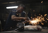 A worker in a dimly lit workshop is focused on welding two metal pieces together, causing bright sparks to fly. The workspace is cluttered with tools and materials, reflecting an industrial setting.