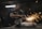 A worker in a dimly lit workshop is focused on welding two metal pieces together, causing bright sparks to fly. The workspace is cluttered with tools and materials, reflecting an industrial setting.
