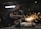 A worker in a dimly lit workshop is focused on welding two metal pieces together, causing bright sparks to fly. The workspace is cluttered with tools and materials, reflecting an industrial setting.