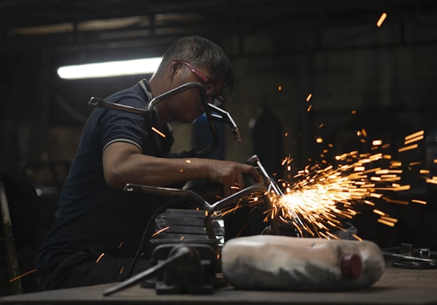A worker in a dimly lit workshop is focused on welding two metal pieces together, causing bright sparks to fly. The workspace is cluttered with tools and materials, reflecting an industrial setting.