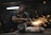A worker in a dimly lit workshop is focused on welding two metal pieces together, causing bright sparks to fly. The workspace is cluttered with tools and materials, reflecting an industrial setting.