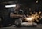 A worker in a dimly lit workshop is focused on welding two metal pieces together, causing bright sparks to fly. The workspace is cluttered with tools and materials, reflecting an industrial setting.