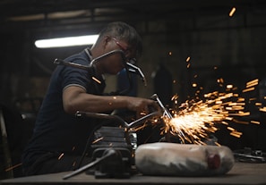 A worker in a dimly lit workshop is focused on welding two metal pieces together, causing bright sparks to fly. The workspace is cluttered with tools and materials, reflecting an industrial setting.